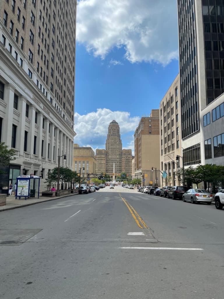 A downtown city street lined with high-rise buildings, leading toward a tall art-deco tower in the distance