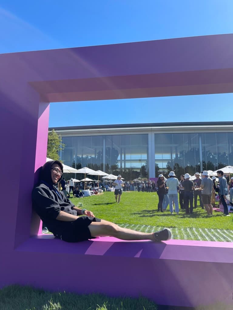 Person sitting in a large purple frame-shaped sculpture on a lawn, with a glass office building and a crowd gathered in the background